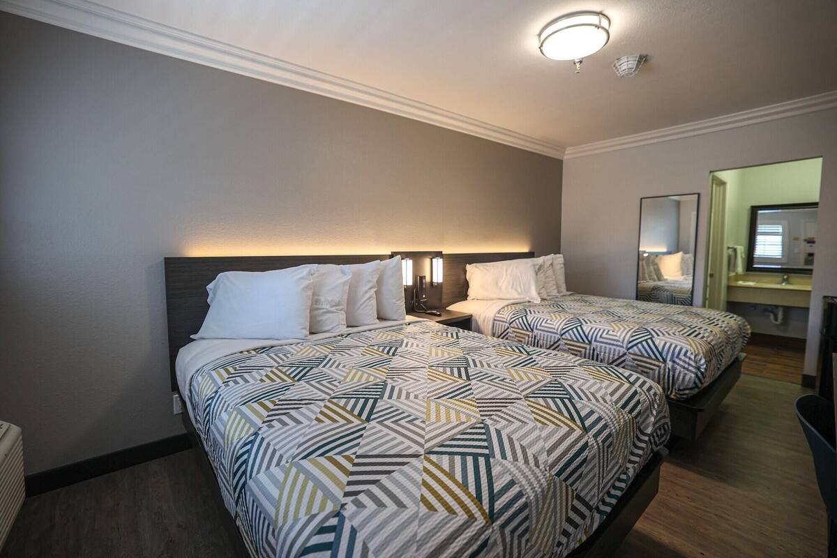 A hotel room with two beds, a nightstand with a lamp, a mirror, and a small desk with a chair.