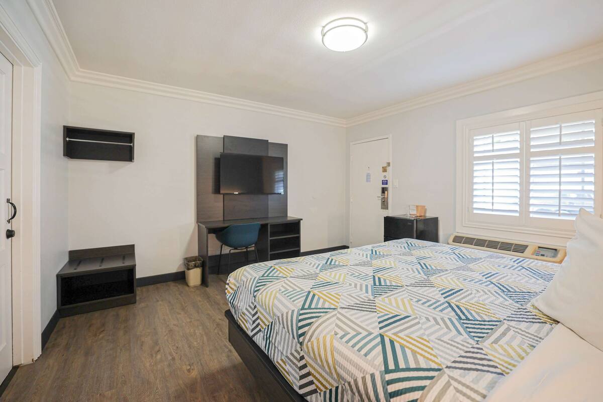A bedroom with a bed, a desk, a chair, a TV, and a window with blinds.