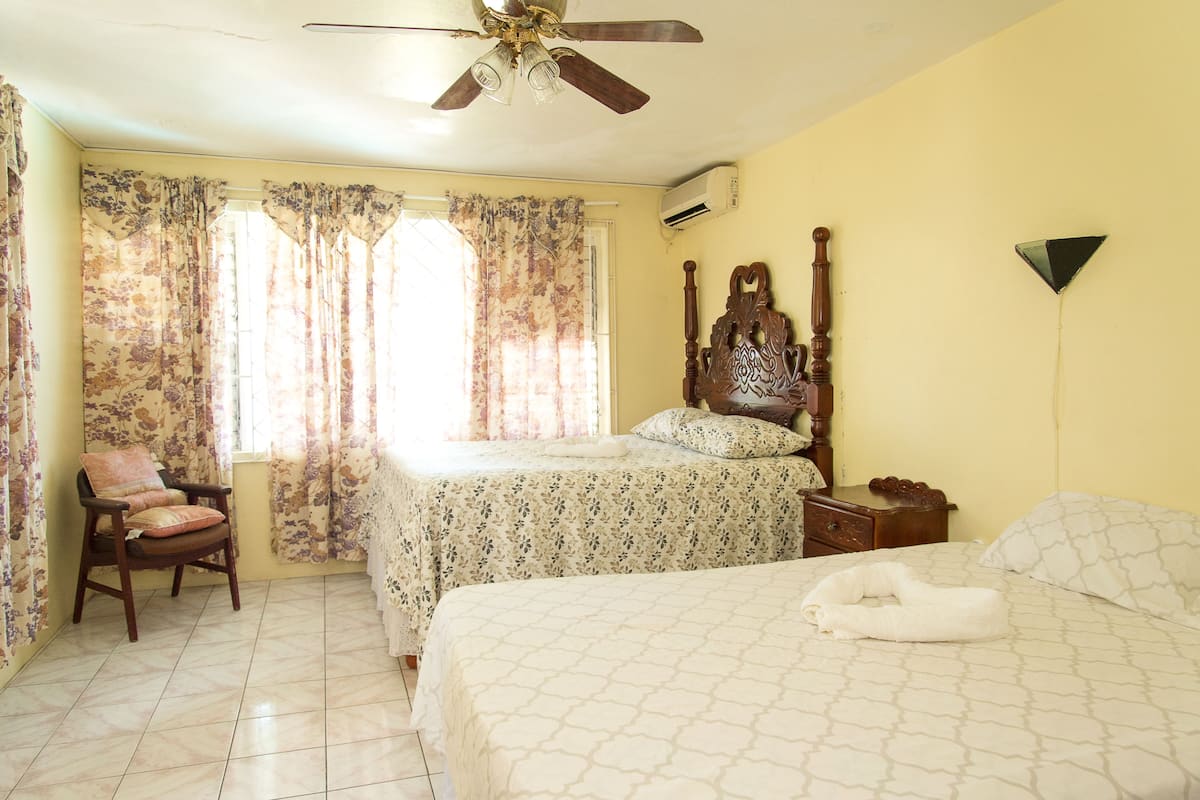 A bedroom with a bed, a chair, a wooden headboard, a ceiling fan, and a window with curtains.