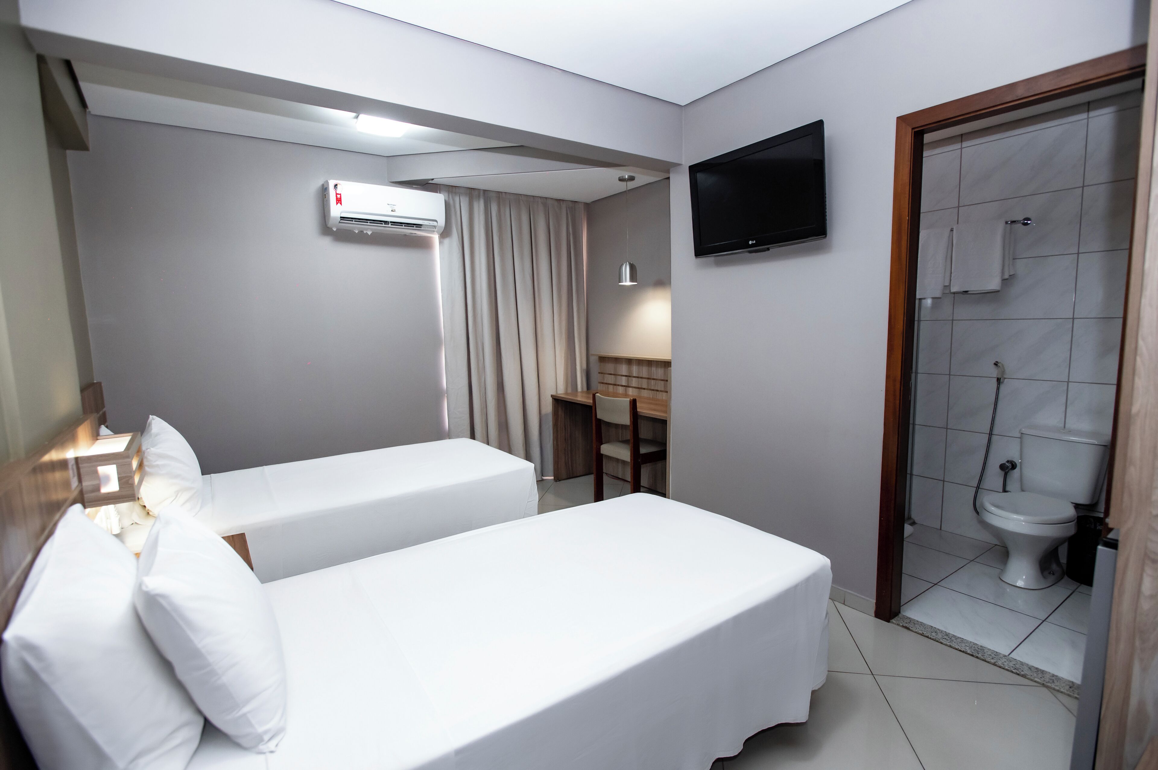 Quarto de hotel com duas camas de solteiro, uma secretária com cadeira, televisão fixada na parede e zona de duche com sanita.
