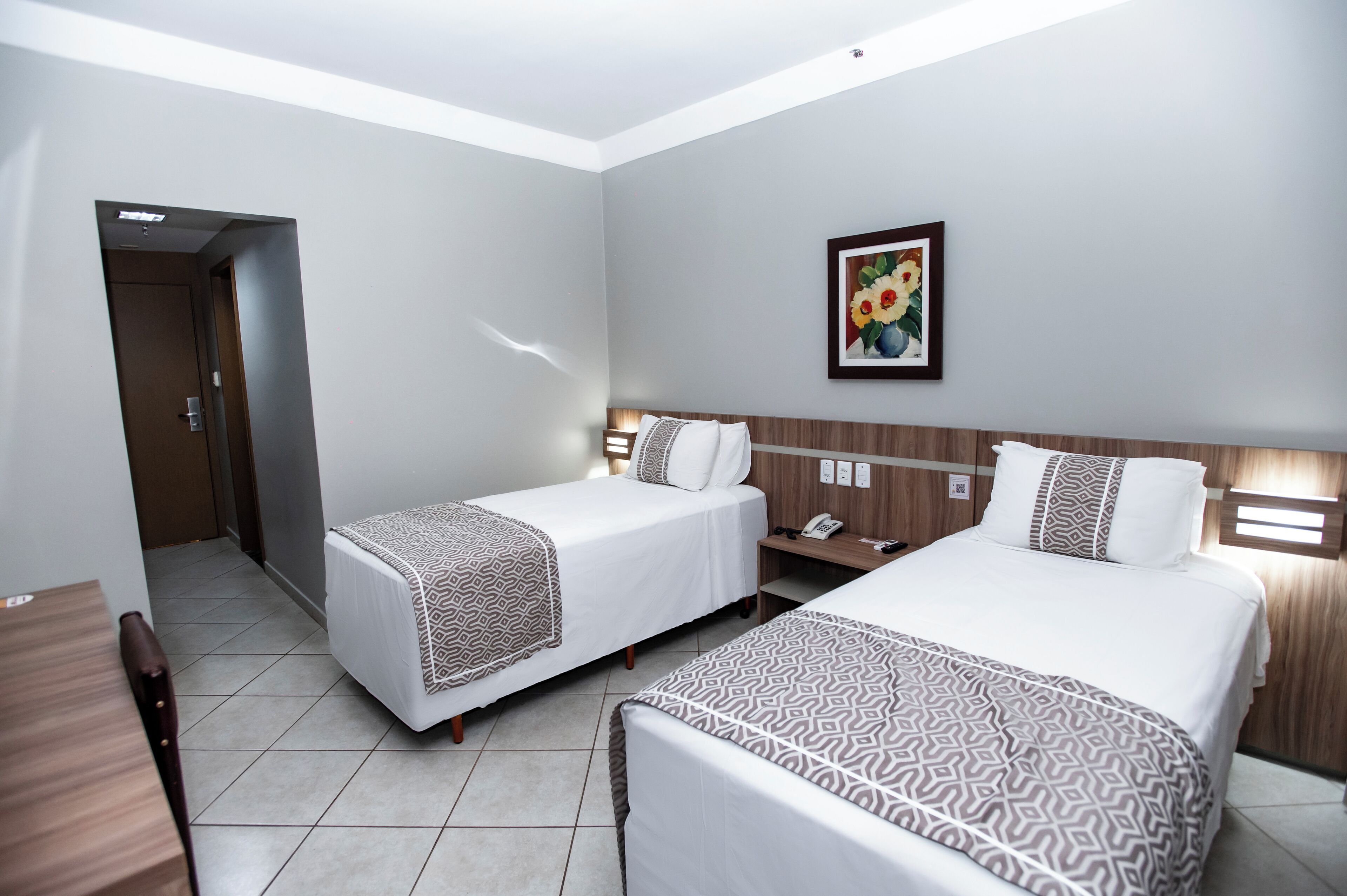 A hotel room with two beds, a wooden headboard, a nightstand, a framed picture on the wall, and a door leading to another room.