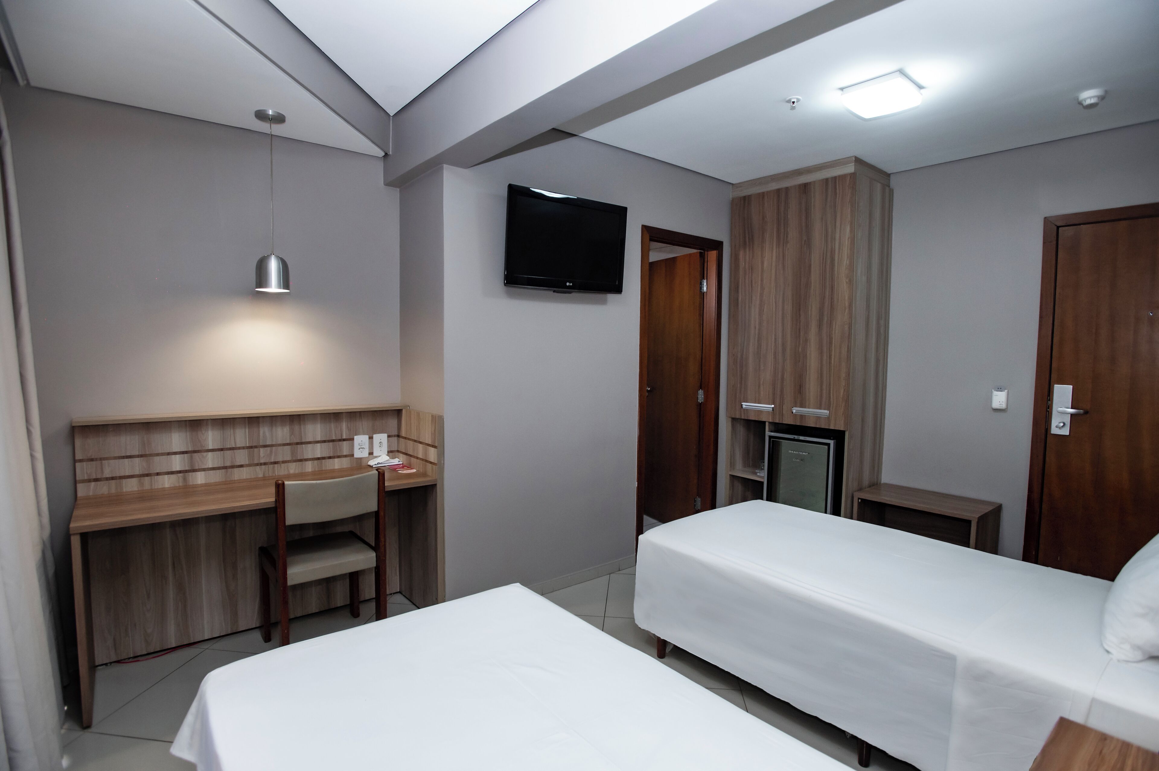 A hotel room with a bed, a desk with a chair, a TV mounted on the wall, and a wardrobe.