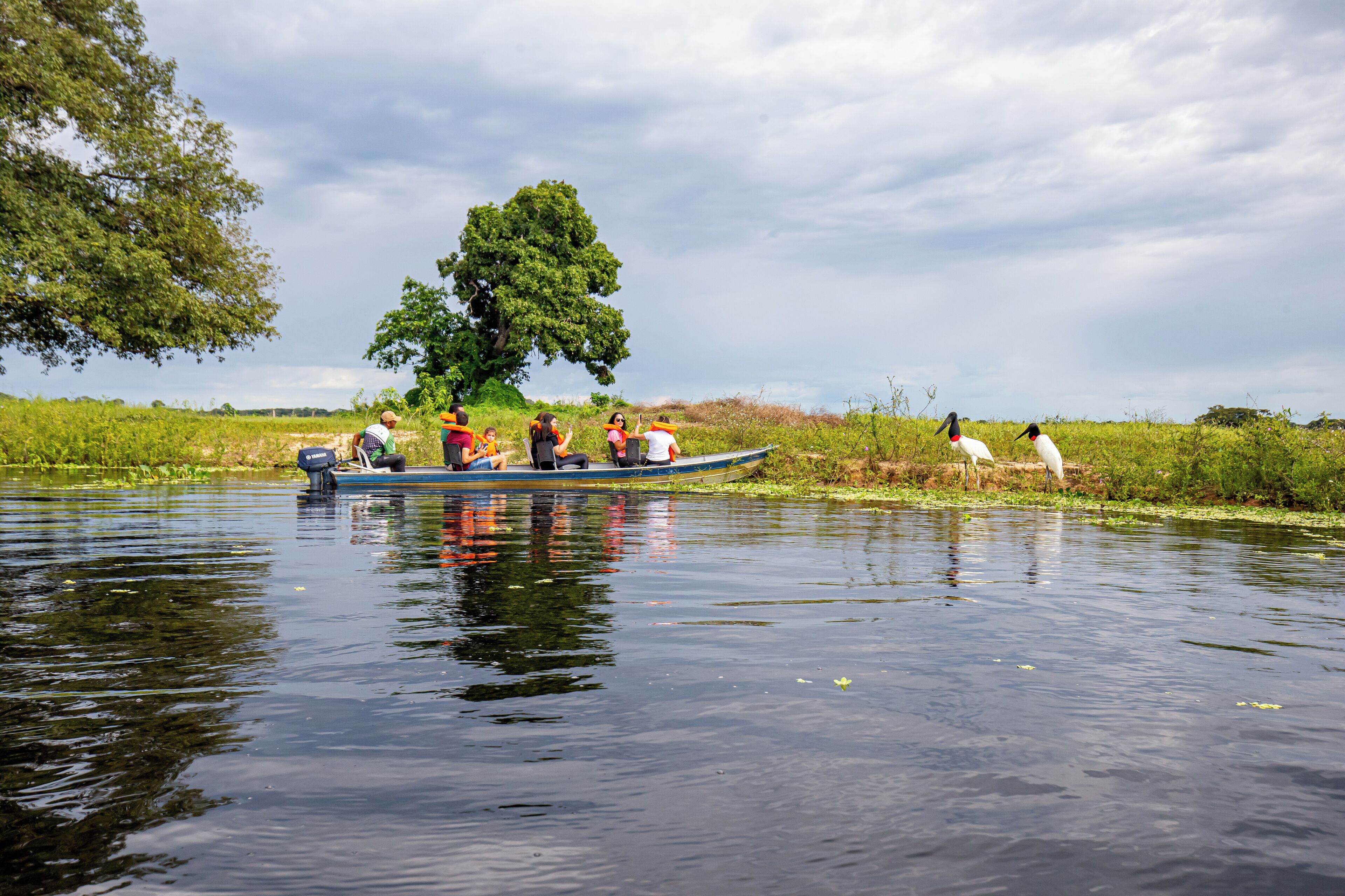 Een boot met passagiers op een rustige rivier, omringd door groen en een bewolkte lucht.