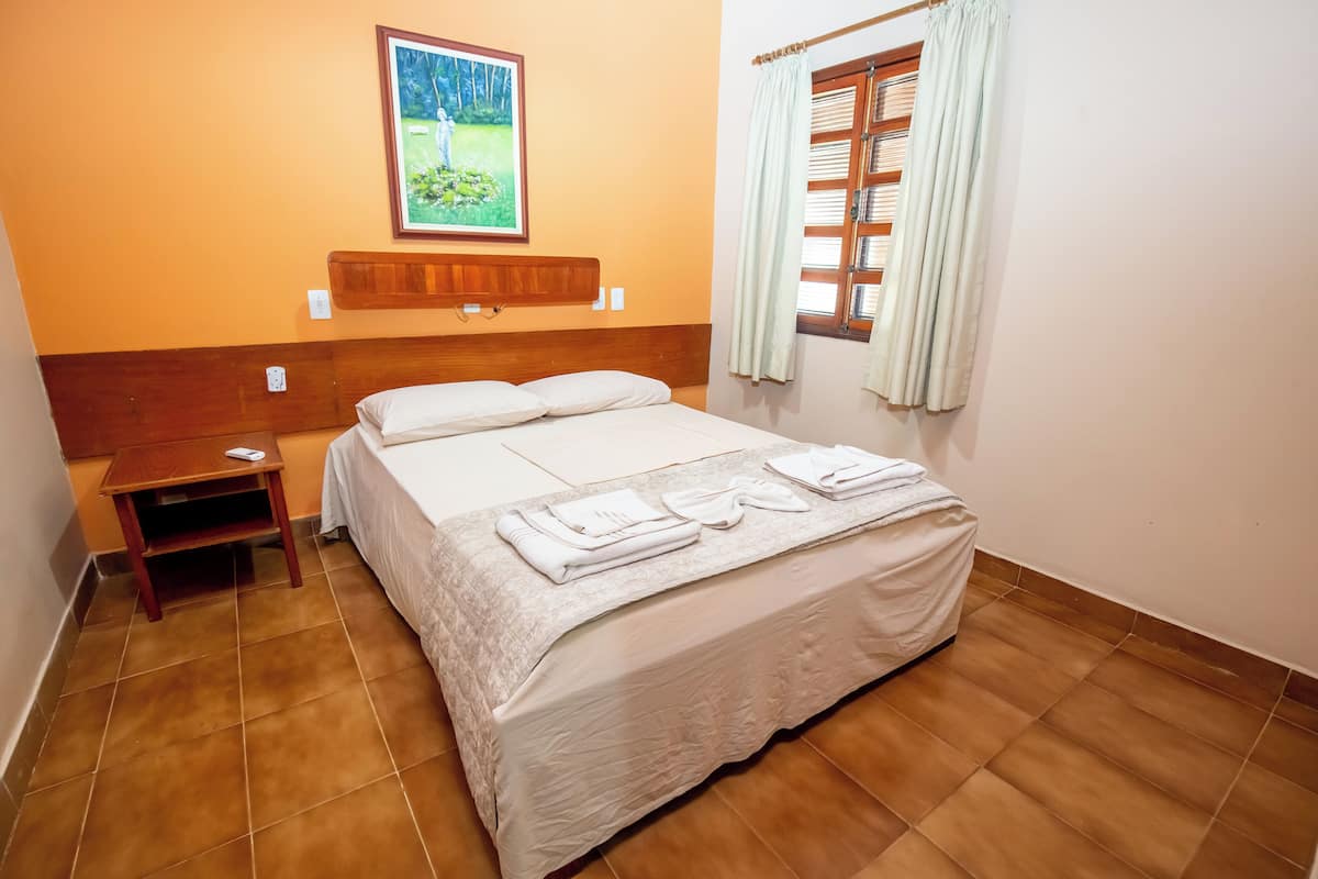 Ein ordentlich bezogenes Bett mit weißen Bettwäsche und Handtüchern, ein Nachttisch aus Holz, ein Bild an der Wand und ein Fenster mit Vorhängen.