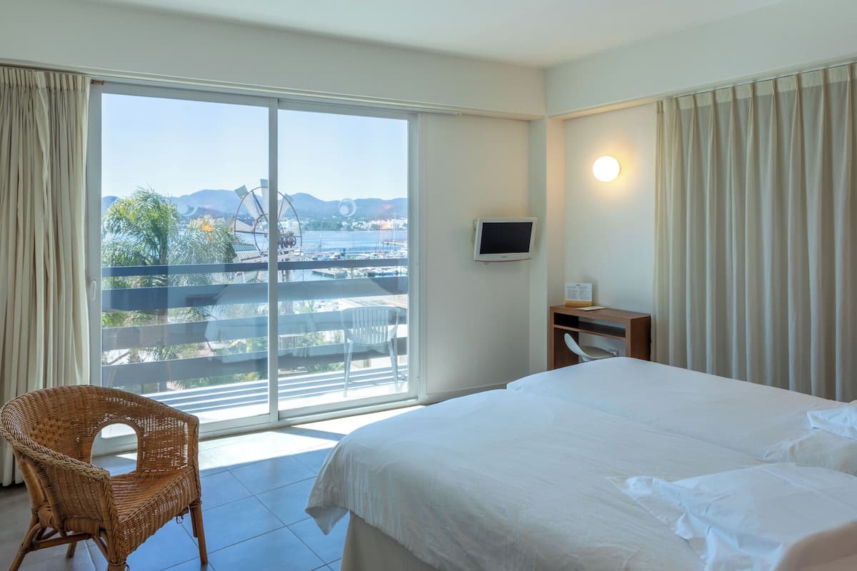 A hotel room with a bed, a chair, a TV, and a balcony with a view of the sea and mountains.