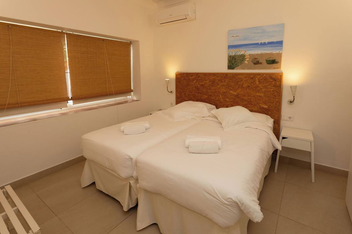 A double bed with two pillows and two towels, a wooden headboard, a small bedside table, a wall-mounted painting of a beach scene, and a window with blinds.