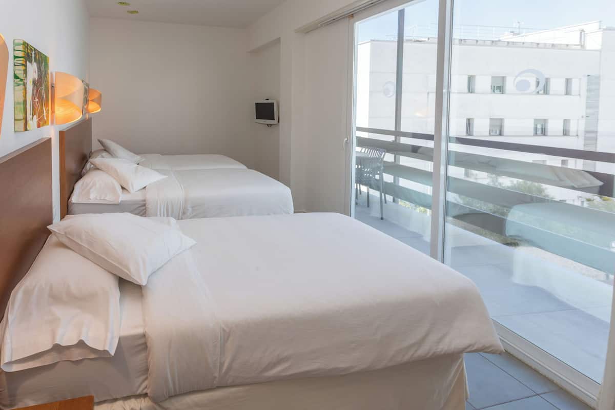 A hotel room with two beds, a TV, and a balcony with a view of buildings.