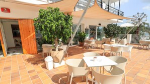 An outdoor patio area with tiled flooring, white tables and chairs, potted plants, and a large umbrella providing shade. - Hotel Es Mitjorn (Sant Antoni de Portmany)