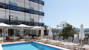 A swimming pool with lounge chairs and umbrellas, a multi-story building with balconies, and a clear sky. - Hotel Es Mitjorn (Sant Antoni de Portmany)