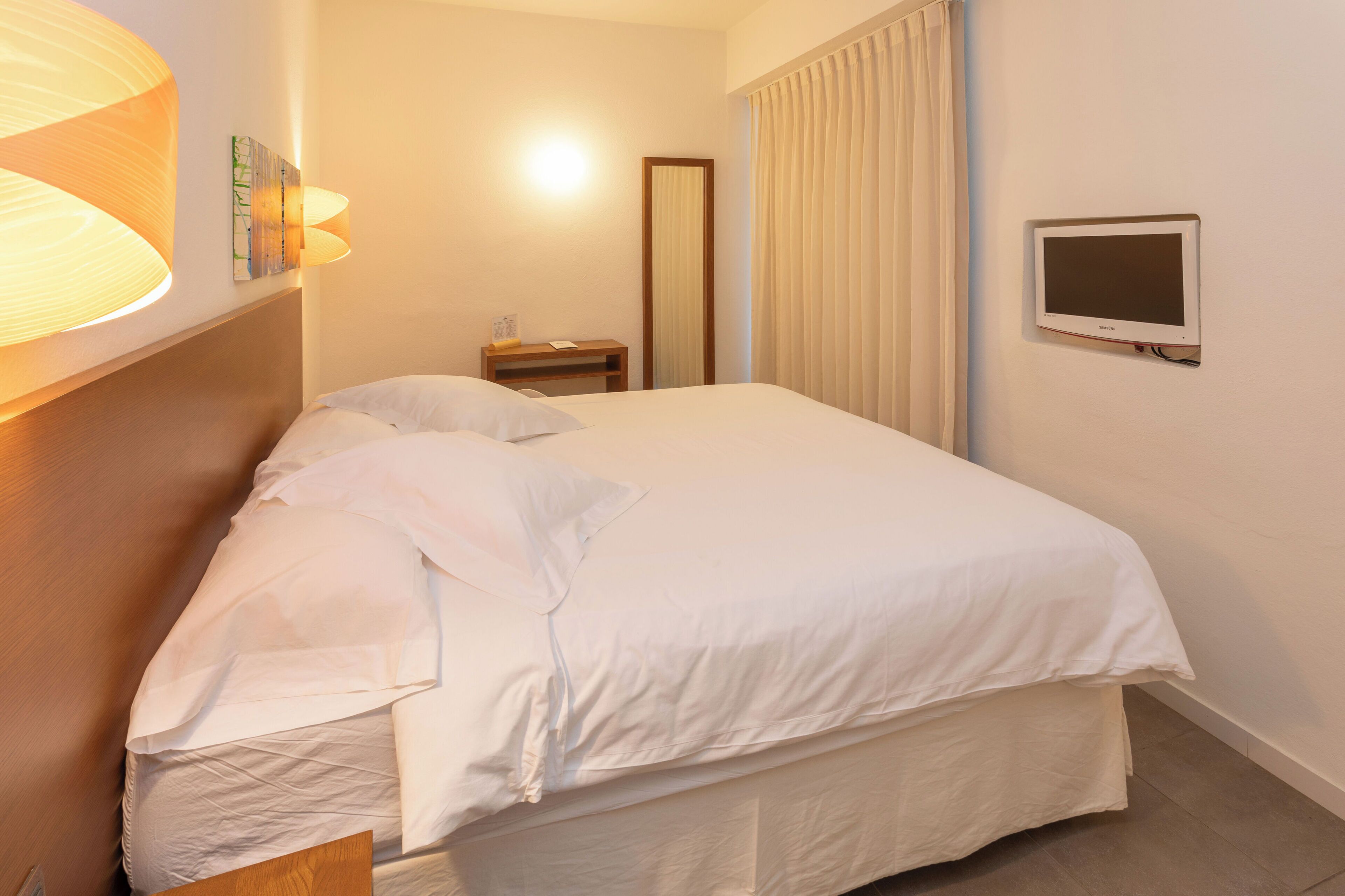 Cama bem arrumada com lençóis brancos, mesa de cabeceira com abajur e televisão fixada na parede.