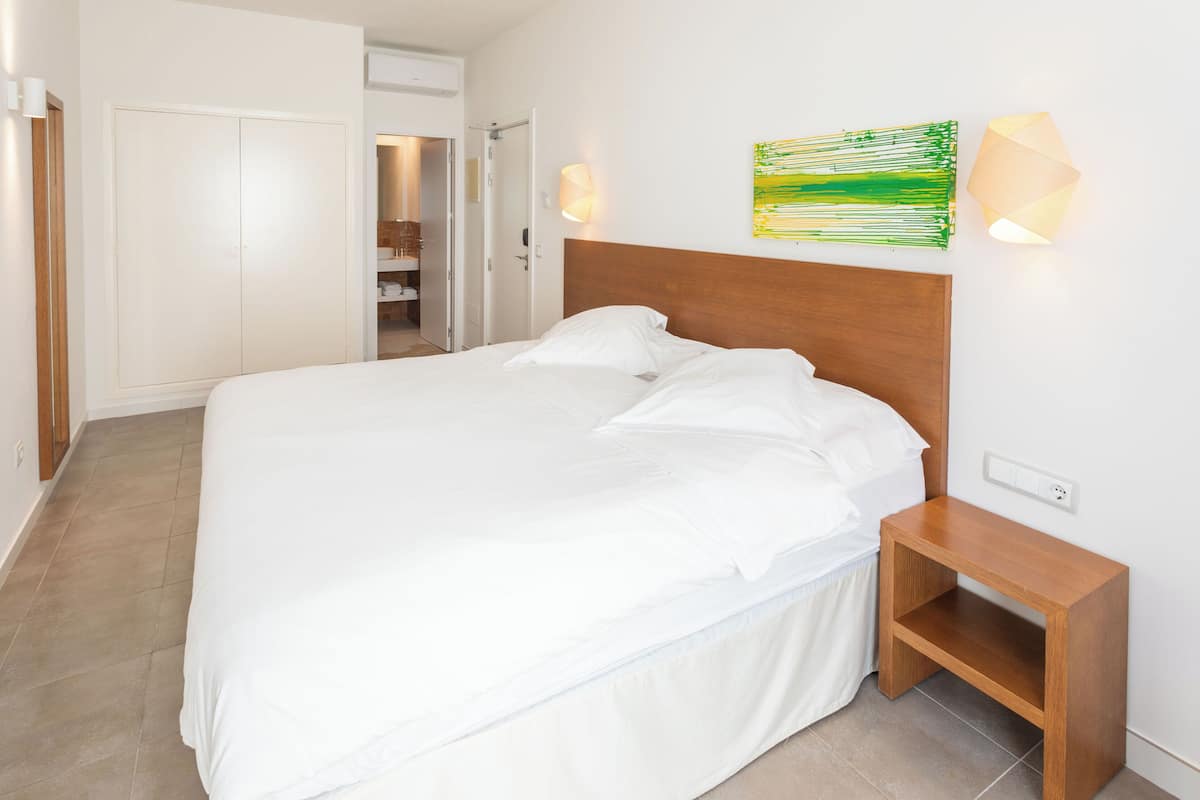A neatly made bed in a hotel room with a wooden headboard, a bedside table, and a wall-mounted lamp.