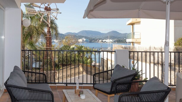 A balcony with wicker furniture, a table, and a view of a body of water and mountains. - Hotel Es Mitjorn (Sant Antoni de Portmany)