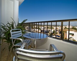 Un balcón con una mesa redonda y dos sillas, con vista a una zona residencial con casas y un cielo azul despejado.