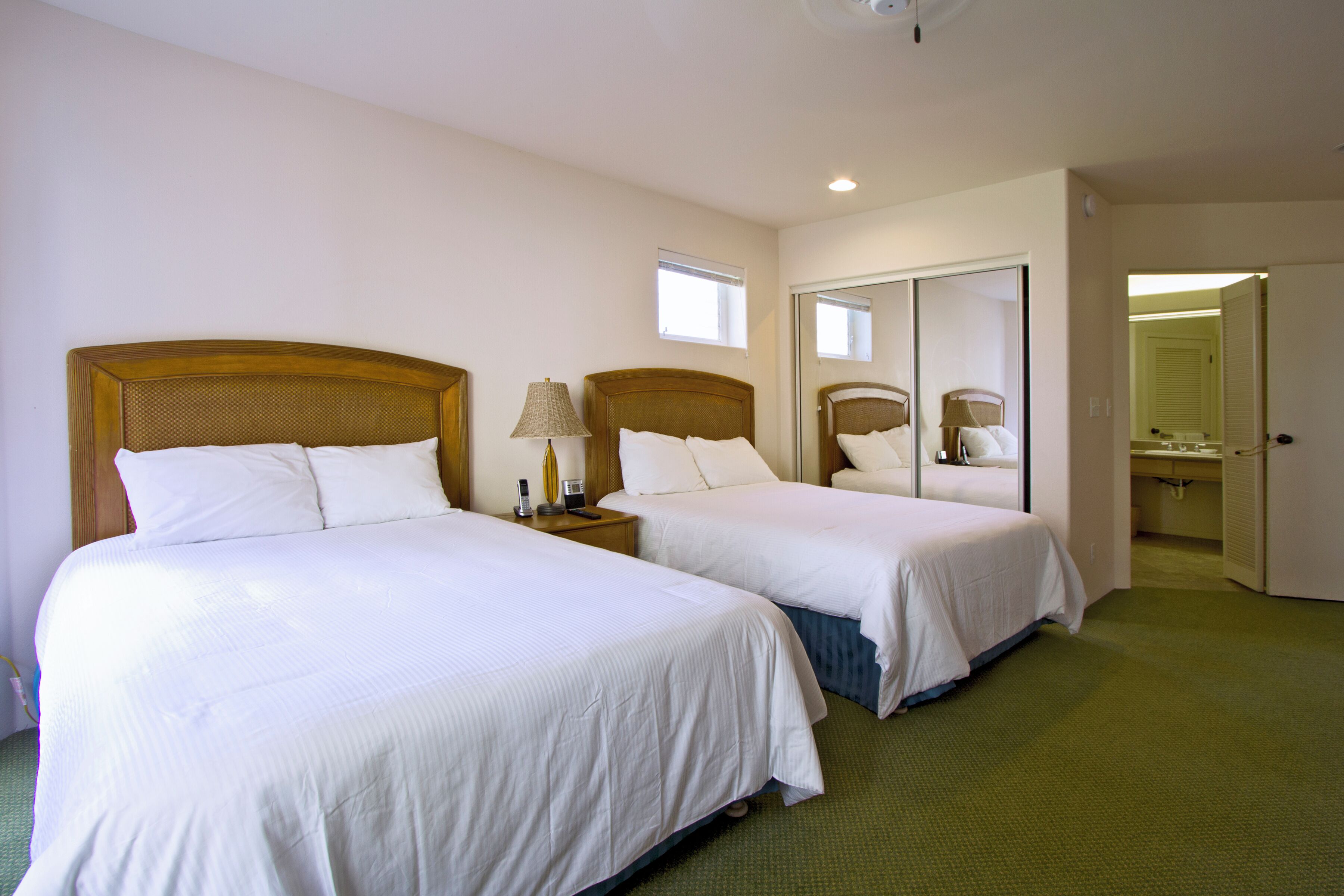 Habitación de hotel con dos camas, una mesita de noche con lámpara, un espejo y un baño visible a través de una puerta abierta.