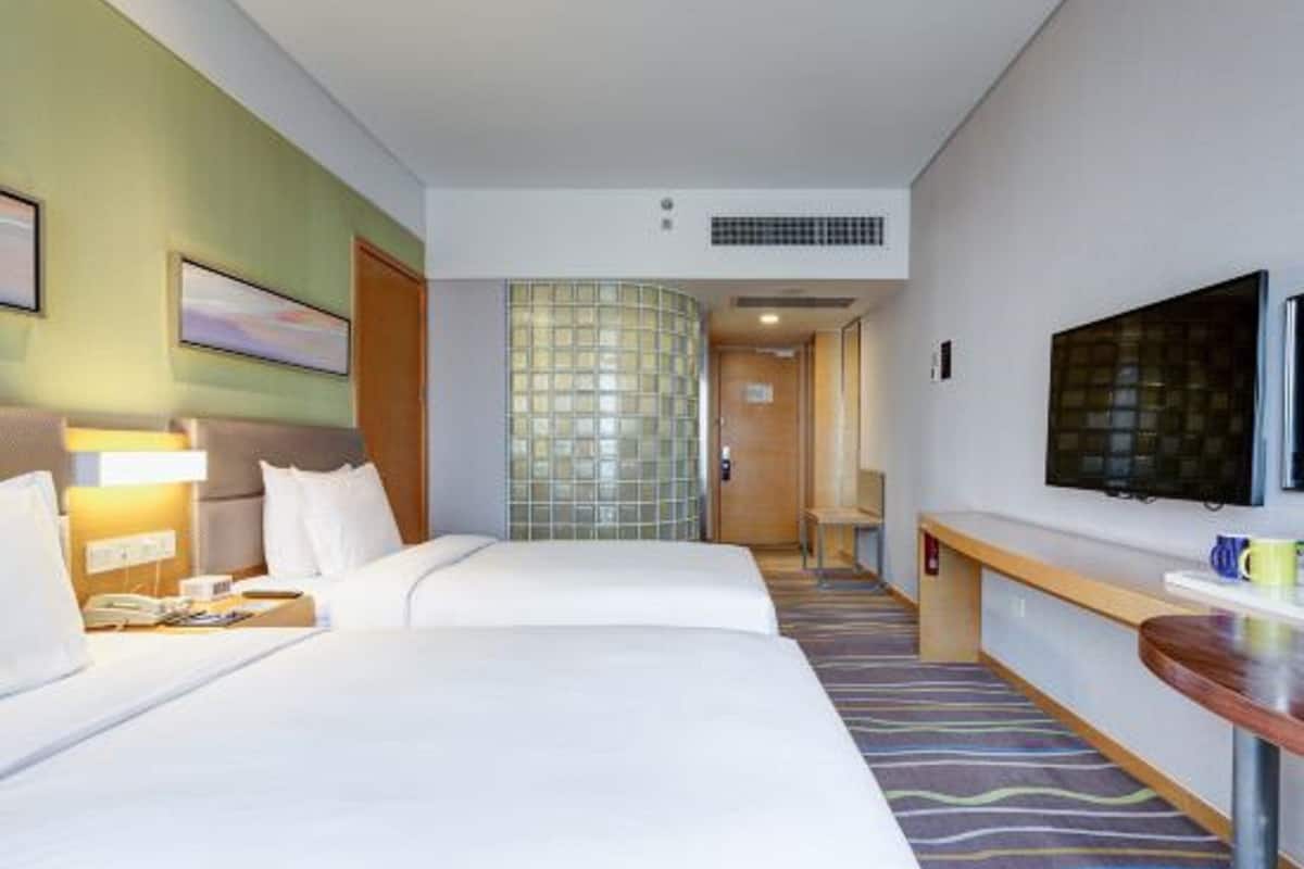 A hotel room with two beds, a television, a desk, and a bathroom visible through a glass door.