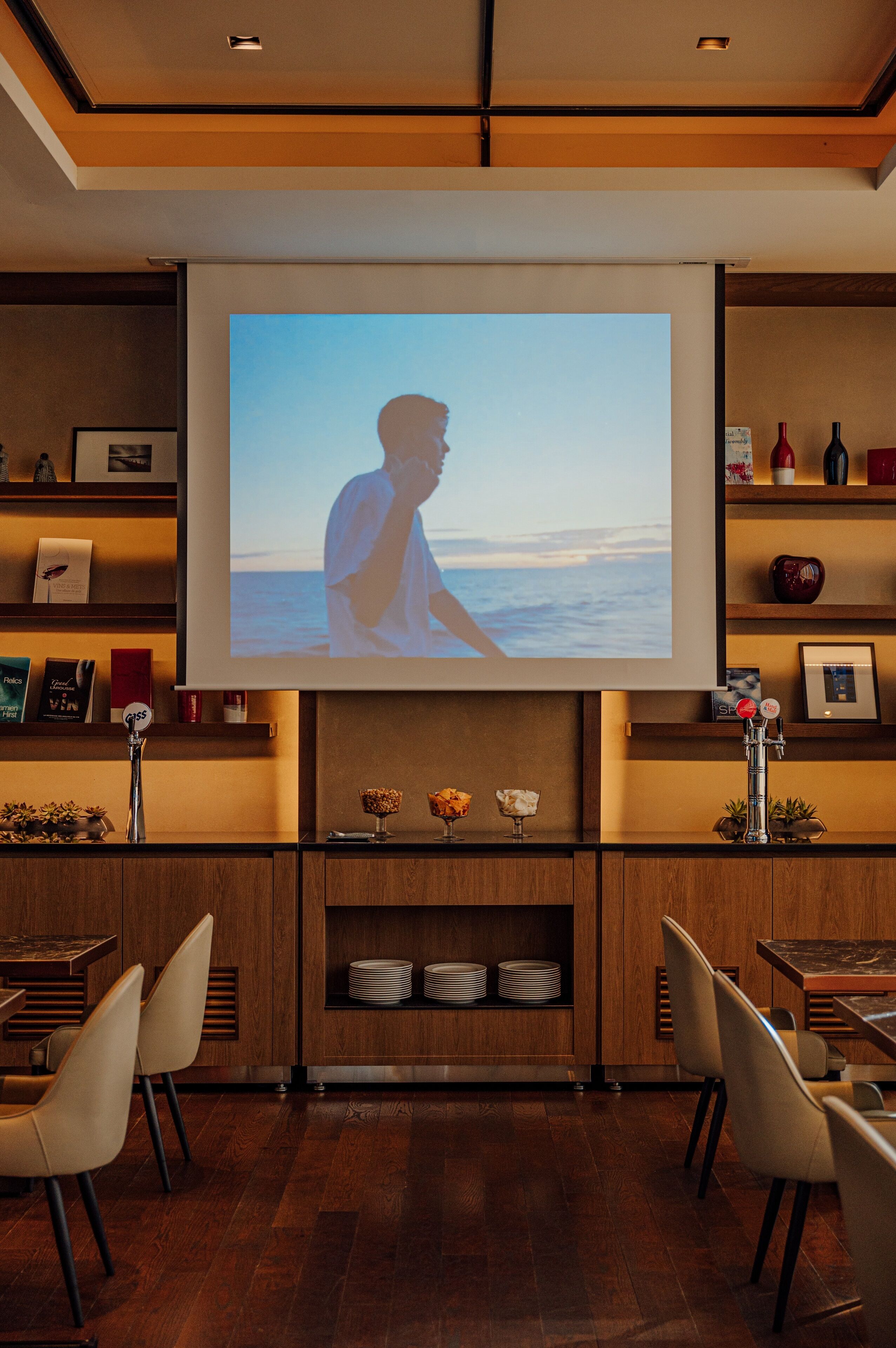 A modern interior with a large screen displaying a beach scene, wooden shelves with books and decorative items, and a dining area with chairs and a table.
