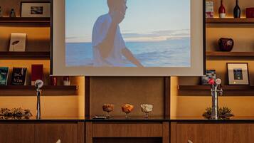 A modern interior with a large screen displaying a beach scene, wooden shelves with books and decorative items, and a dining area with chairs and a table.