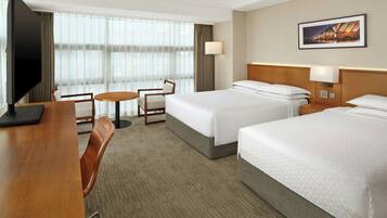 A hotel room with two beds, a desk, a chair, a TV, and a large window with curtains.