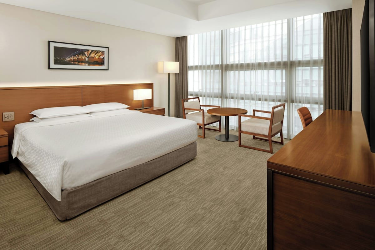 A hotel room with a large bed, a desk, two chairs, a lamp, and a large window with curtains.