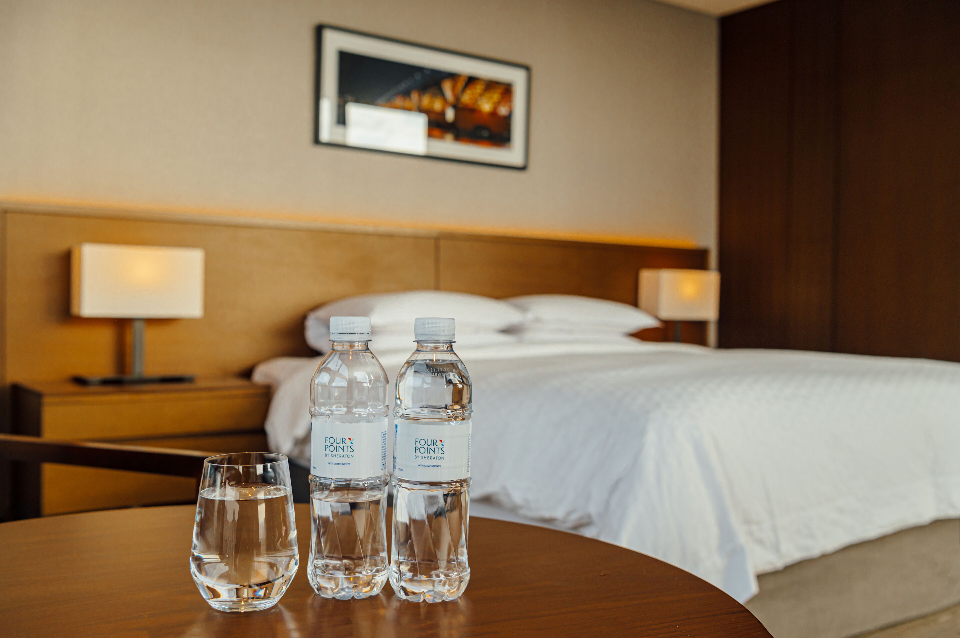 A hotel room with a neatly made bed, two bedside lamps, and three bottles of water on a wooden table.