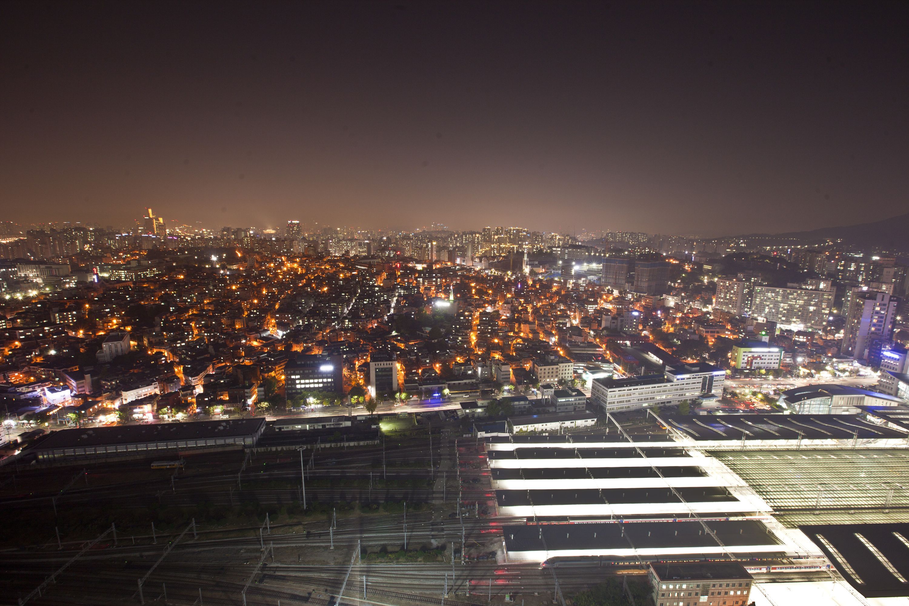 Un paisaje urbano nocturno con edificios iluminados y una amplia zona abierta con estructuras que parecen parte de un centro de transporte.