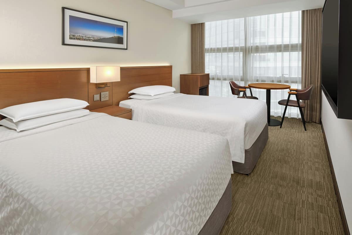 A hotel room with two beds, a desk with a chair, a TV, and a large window.