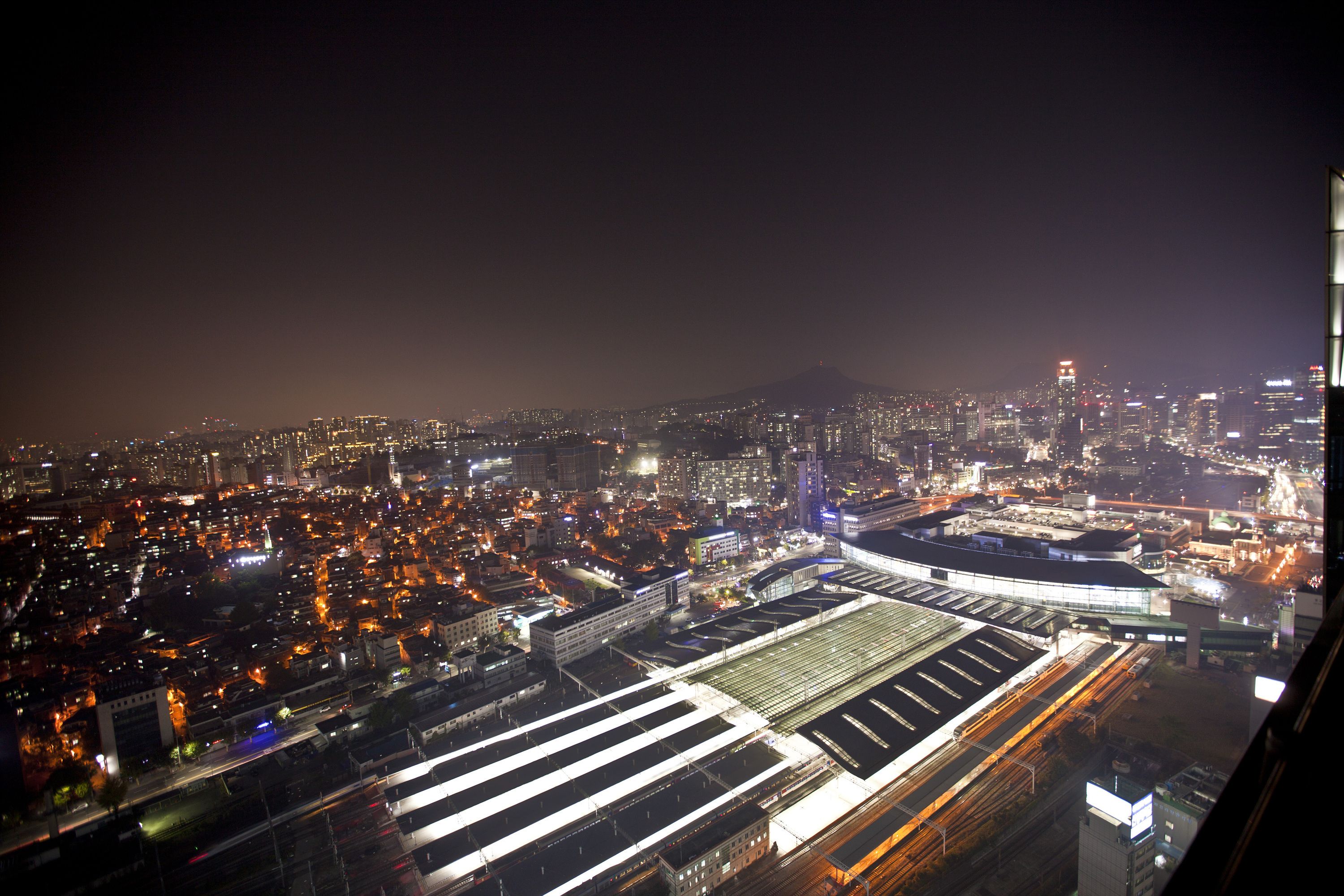 Un paisaje urbano nocturno con una estación de tren destacada y zonas urbanas circundantes.