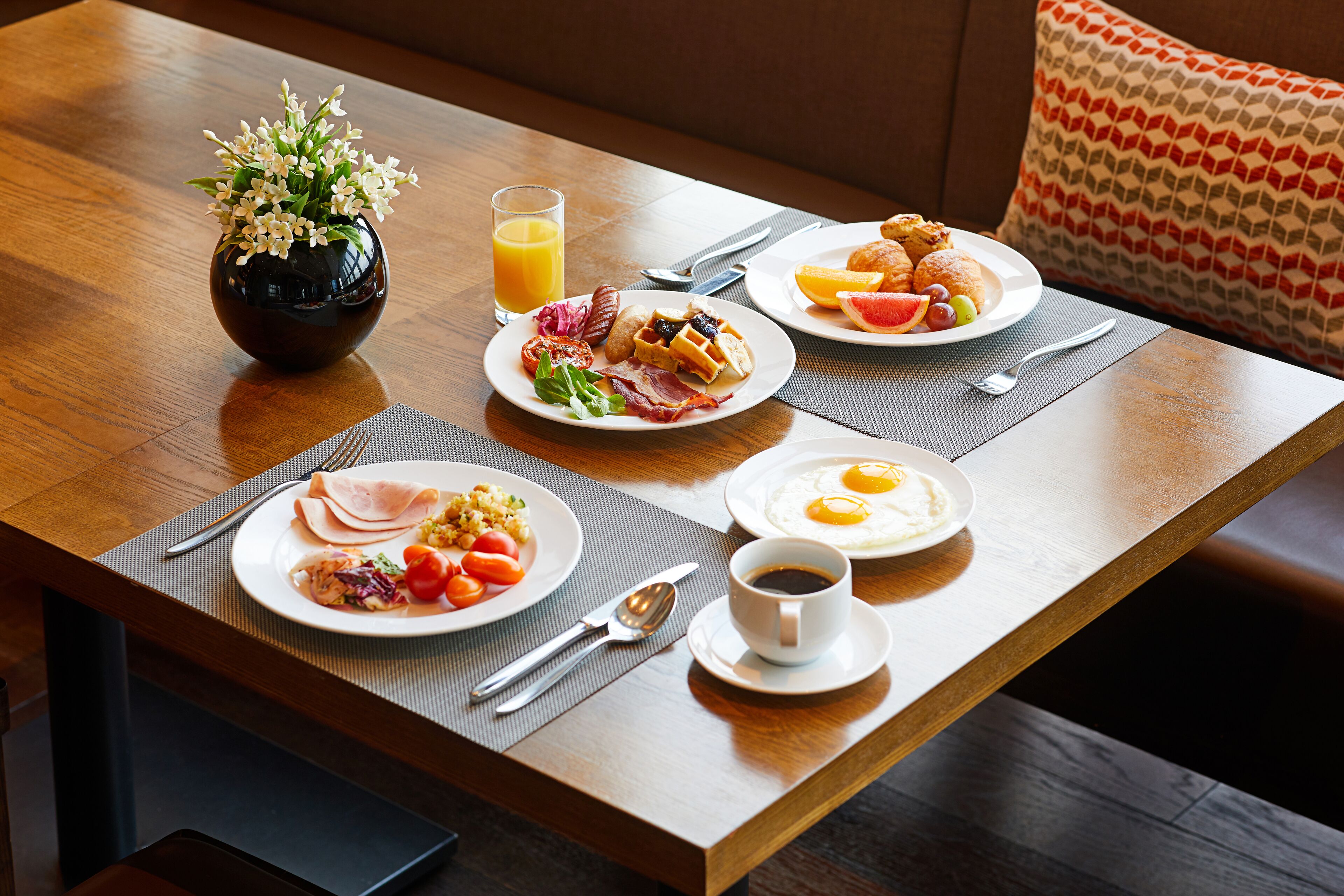 Una mesa de desayuno con platos de comida, un florero de flores y un vaso de jugo de naranja.