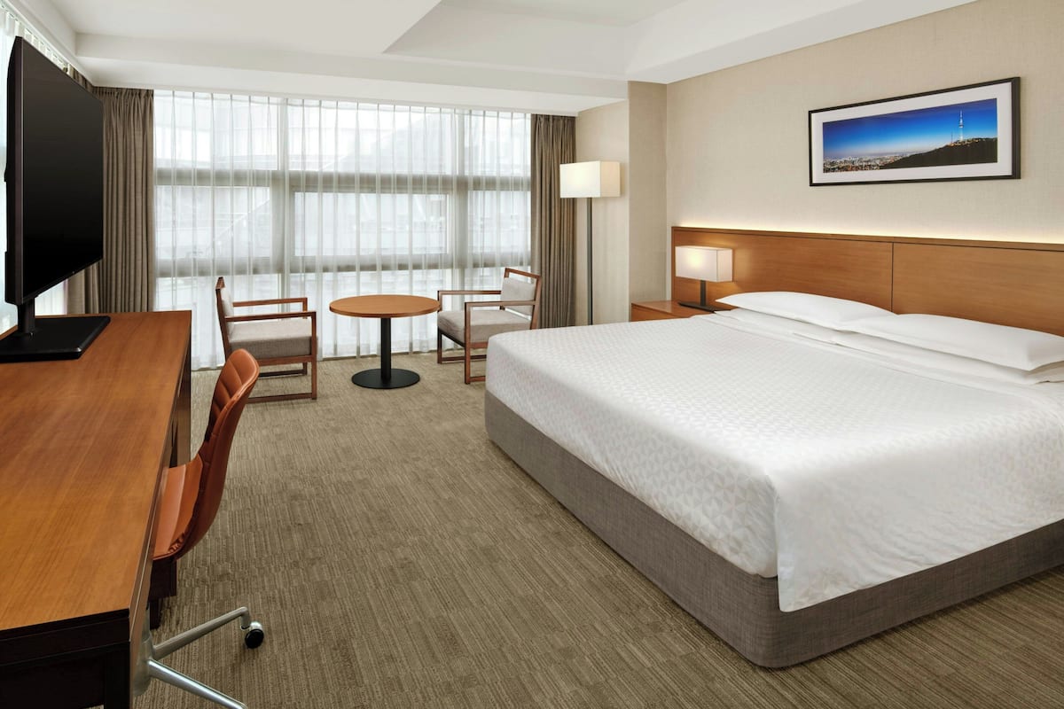 A hotel room with a bed, a desk, a chair, a small table, and a TV.