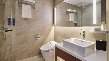 Un baño moderno con ducha, inodoro y lavabo, que cuenta con un espejo grande y baldosas color crema.