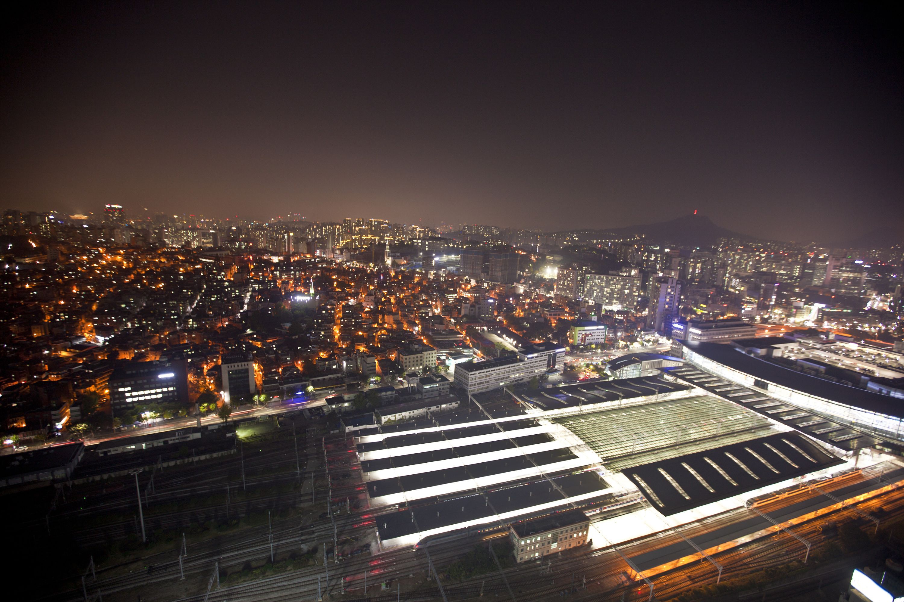 Un paisaje urbano nocturno con edificios iluminados y una amplia zona con una cuadrícula de vías, probablemente una estación de tren.