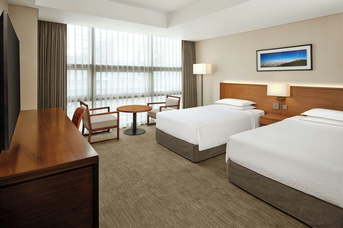 A hotel room with two beds, a desk, a chair, a round table, and a large window with curtains.