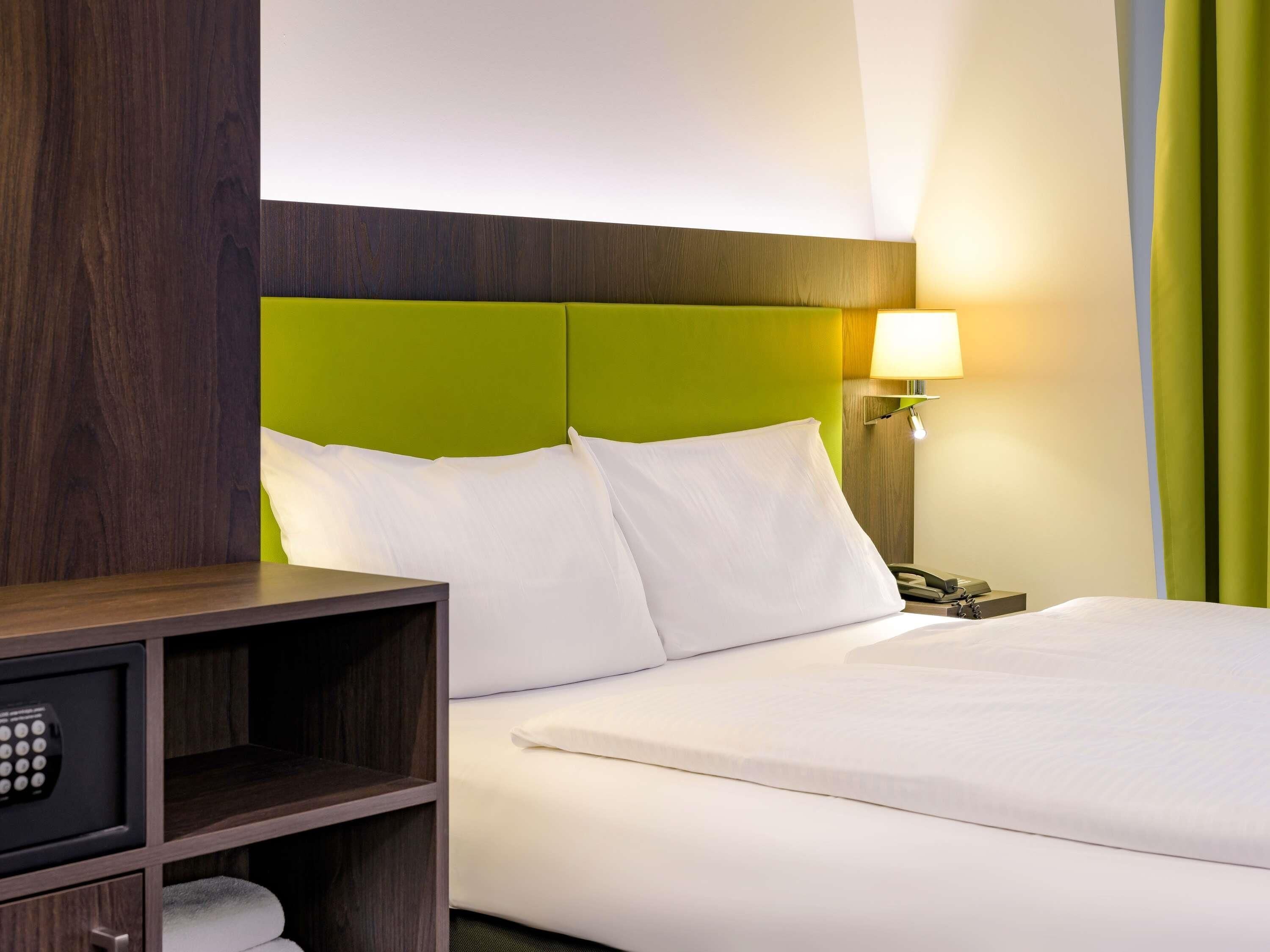 Hotellihuone, jossa on vihreä pääty, yöpöytälamppu, puhelin ja minibaari.