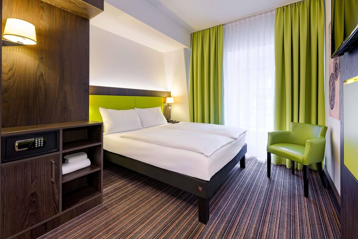 A hotel room with a large bed, a green chair, a desk with a telephone, and a television on the wall.