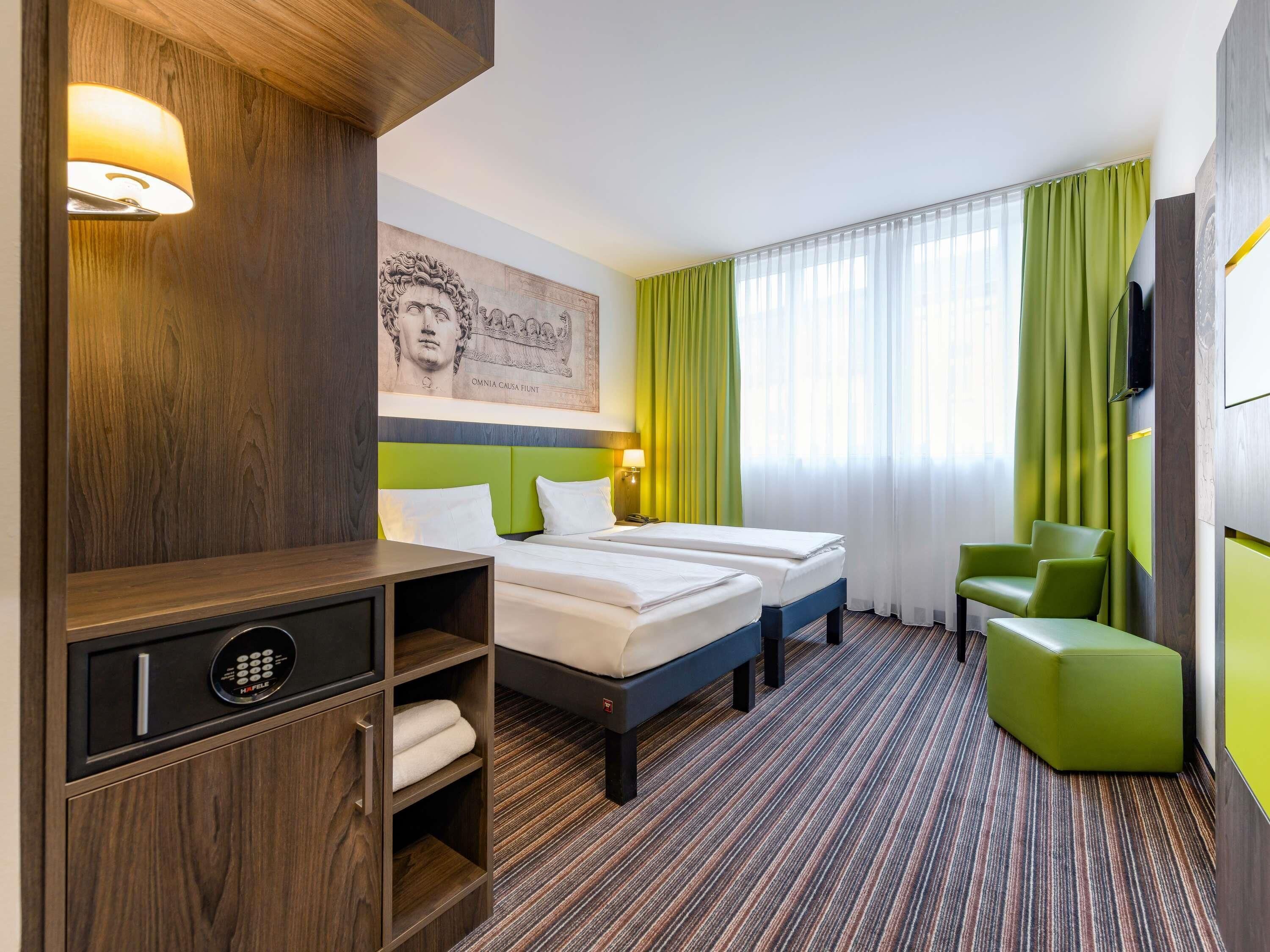 Habitación de hotel con dos camas, un mueble de madera con minibar, una silla verde y un ventanal con cortinas verdes.