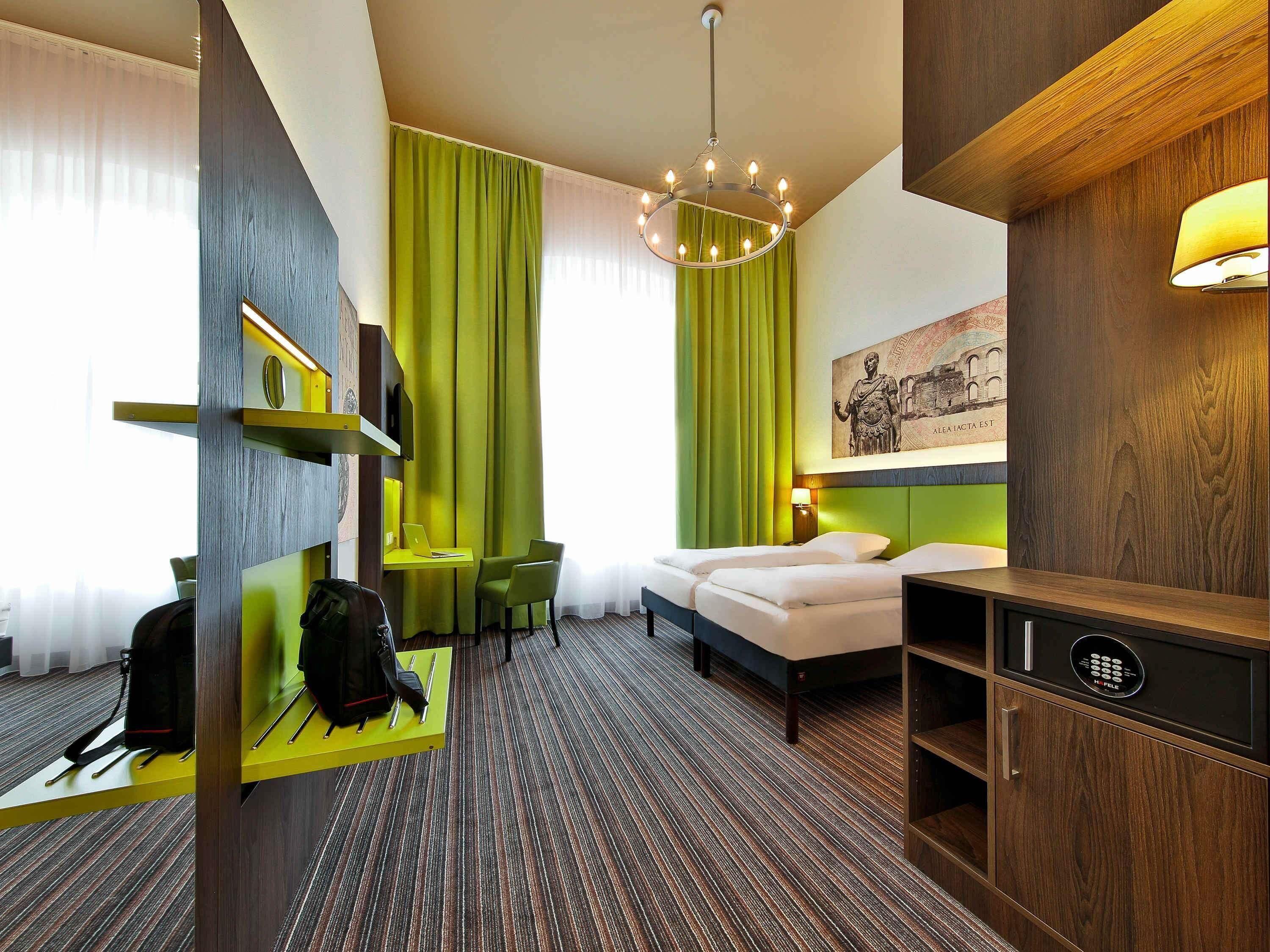 A hotel room with a bed, a desk with a laptop, a chair, a green curtain, a chandelier, and a painting on the wall.