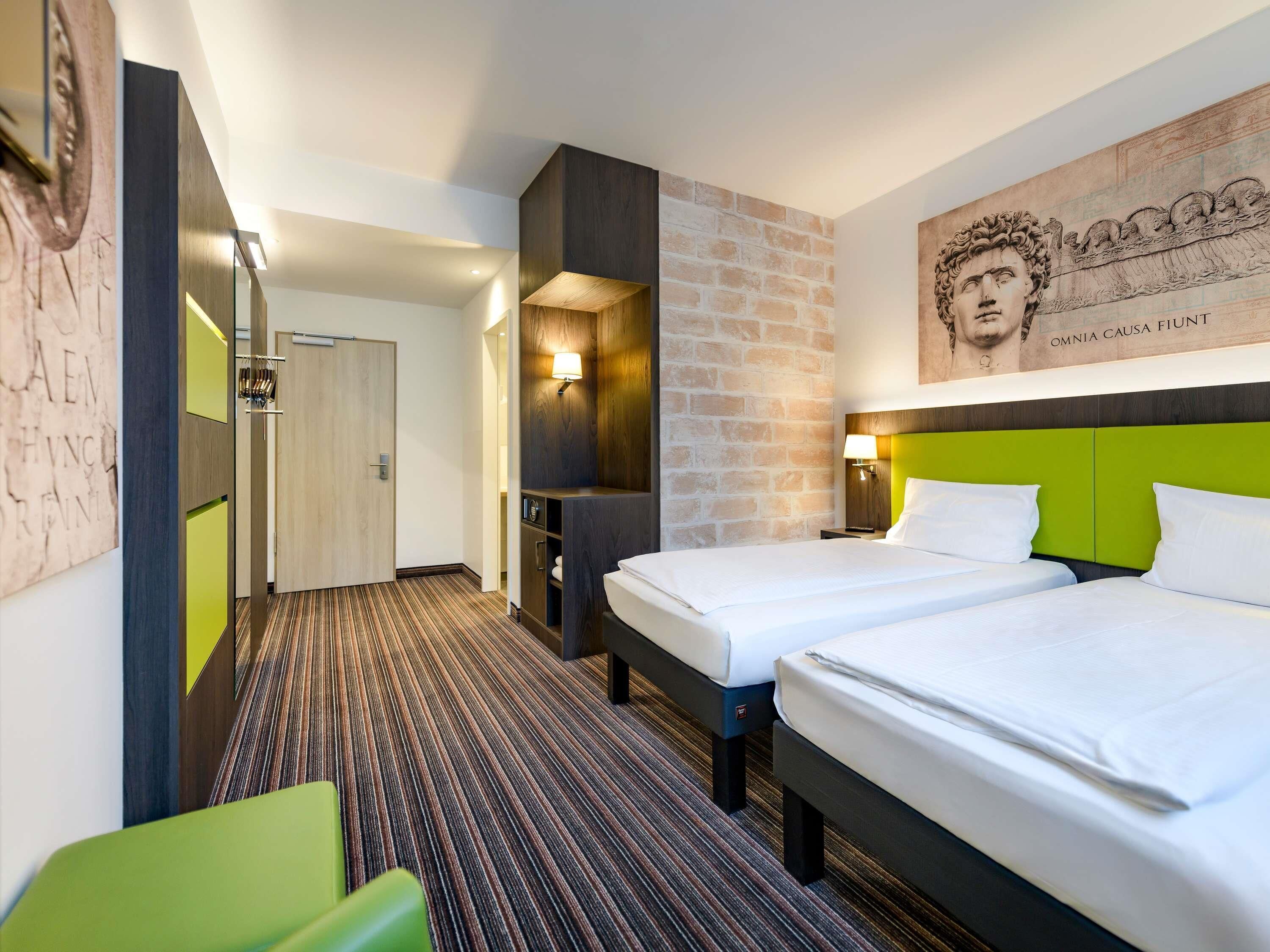 Une chambre d’hôtel avec deux lits, un mur décoratif en briques, une tête de lit verte et une grande œuvre d’art au-dessus du lit.