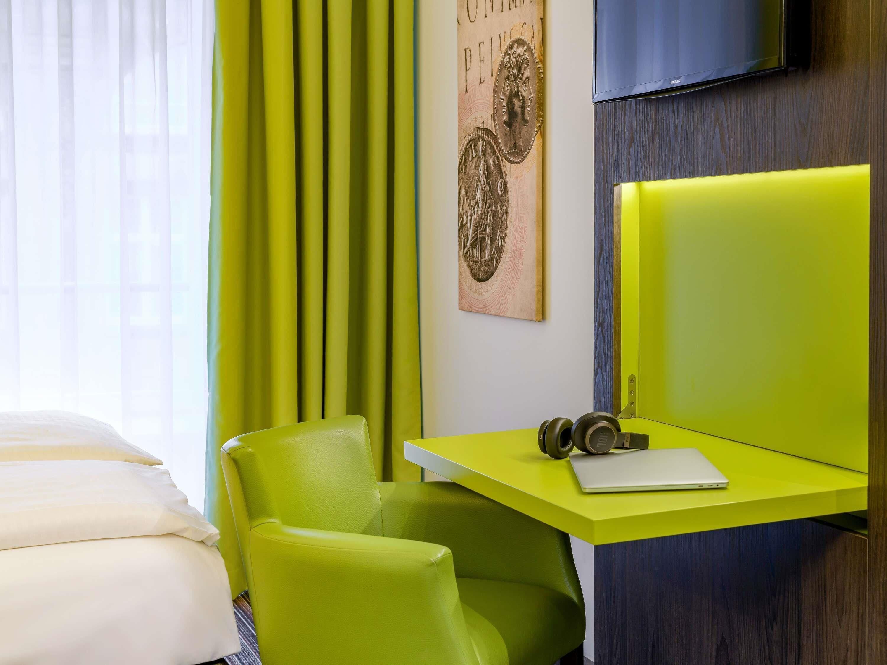 Een hotelkamer met een bed, een groene fauteuil, een bureau met een hoofdtelefoon en een laptop, en een televisie aan de muur.