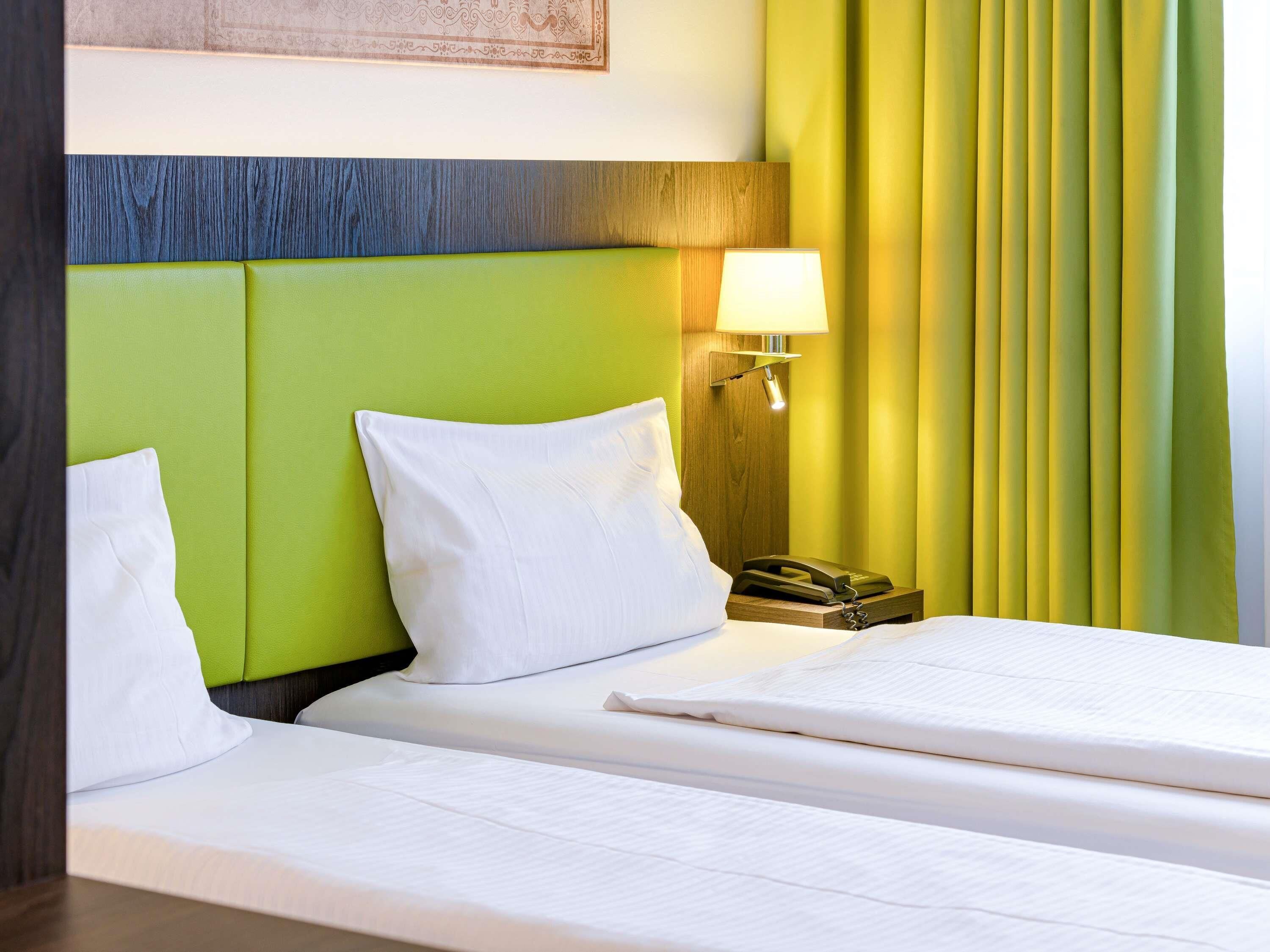Habitación de hotel con cabecera verde, ropa de cama blanca, lámpara de noche y teléfono en la mesita.