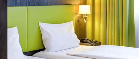 Habitación de hotel con cabecera verde, ropa de cama blanca, lámpara de noche y teléfono en la mesita.
