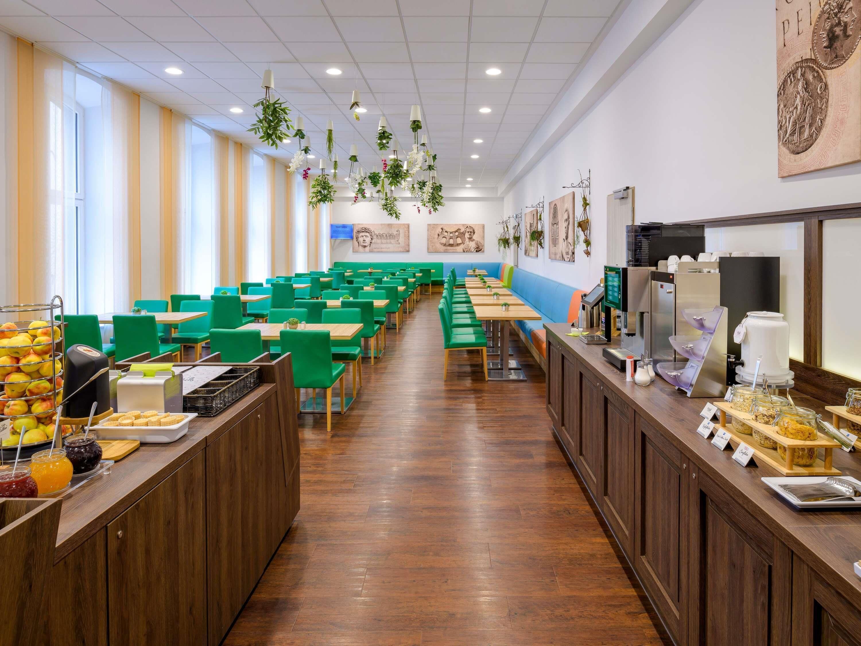 Una cafetería con sillas verdes, mesas de madera y una zona de bufé con comida y bebidas.