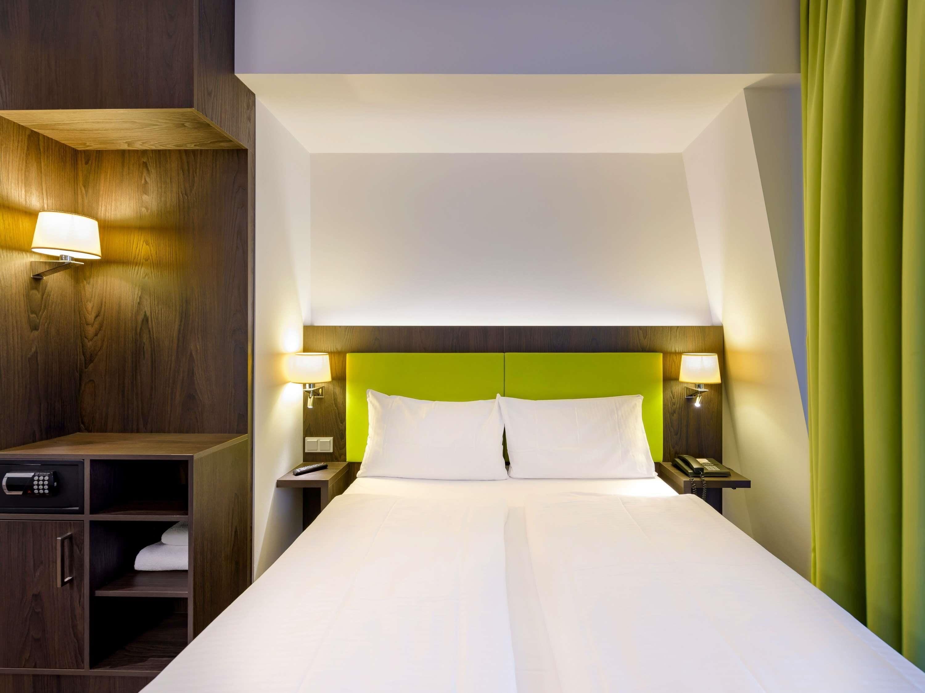 A hotel room with a large bed, two bedside tables with lamps, a wooden wardrobe, and a green curtain.