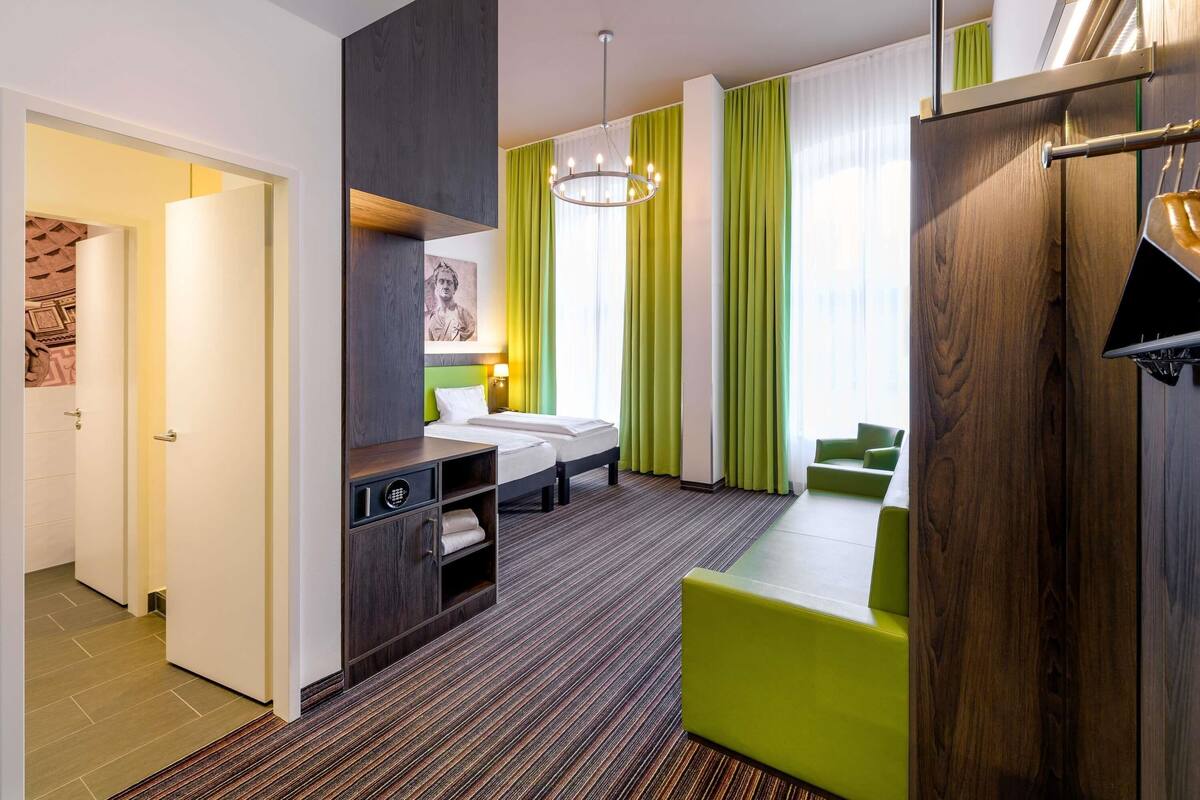 A hotel room with a bed, a desk, a green chair, a wardrobe, and a view of the city.