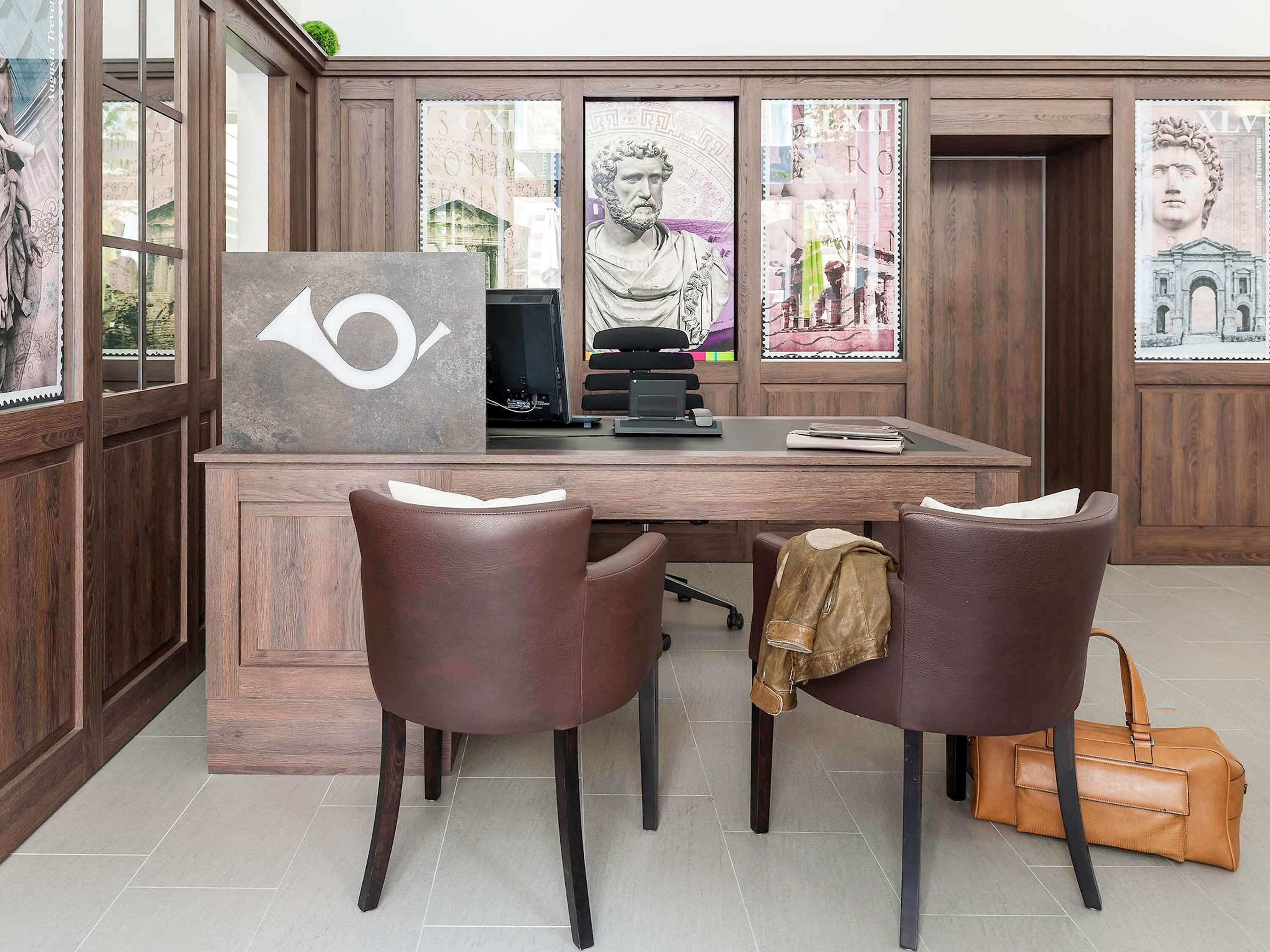 A wooden desk with a computer, two leather chairs, a brown leather bag, and framed artwork on the walls.