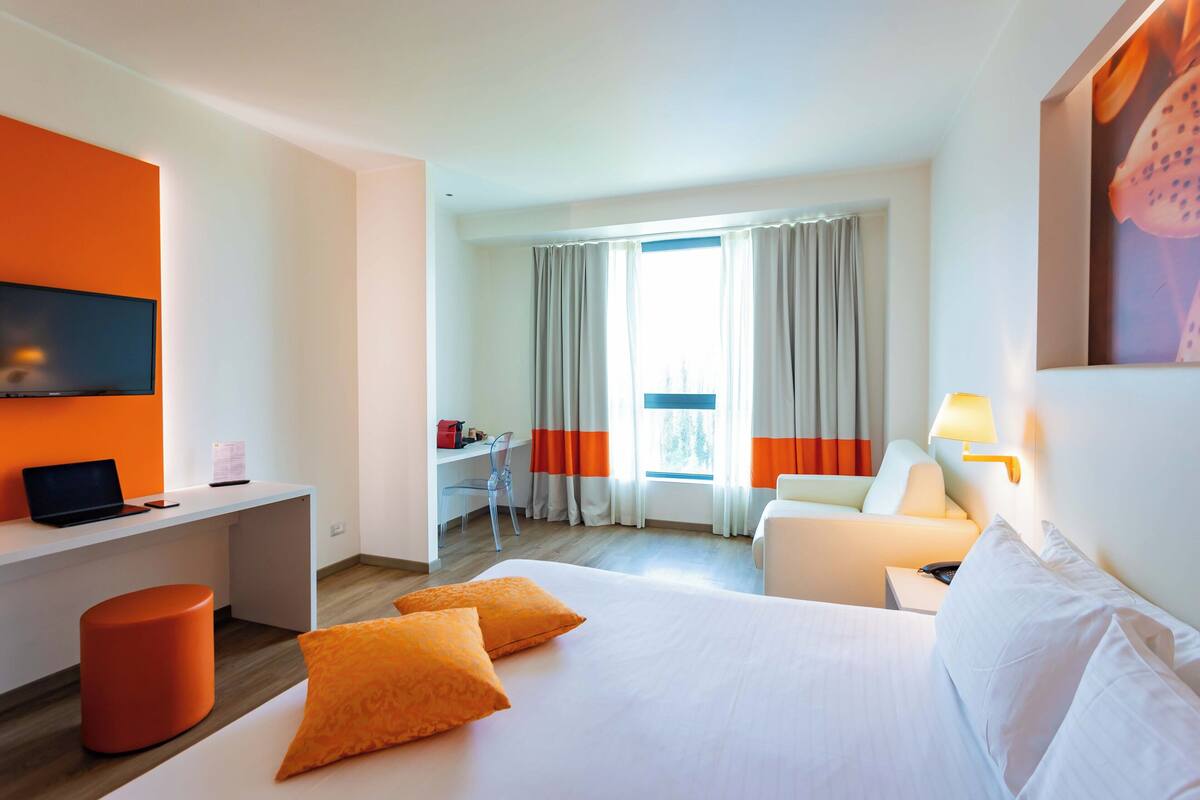 Une chambre d’hôtel avec un lit, un bureau, une chaise, une télévision et une fenêtre avec des rideaux.