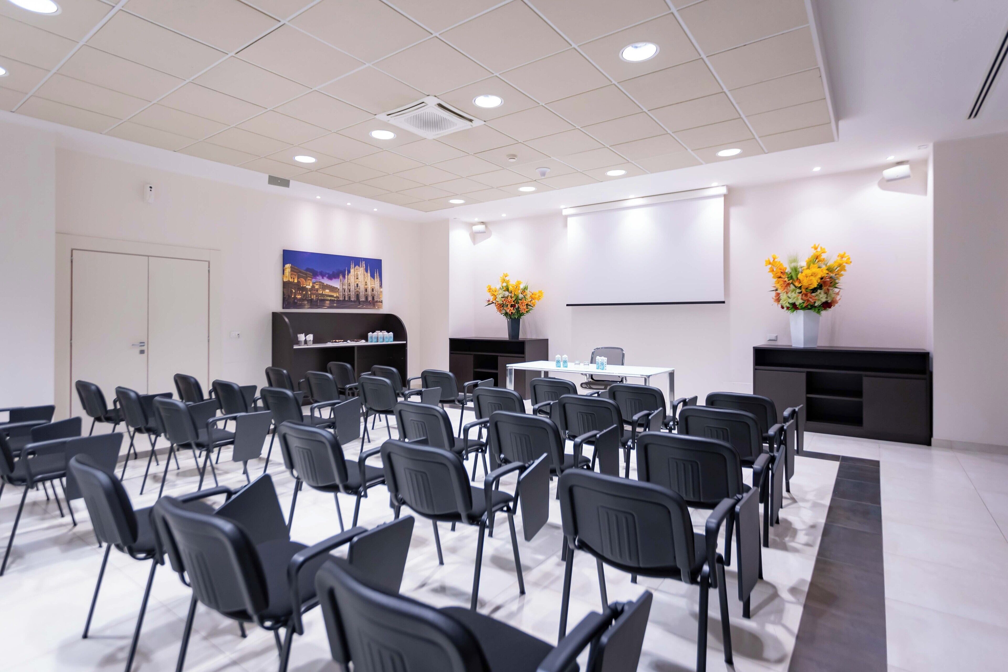 A conference room with rows of chairs, a whiteboard, and two flower arrangements.