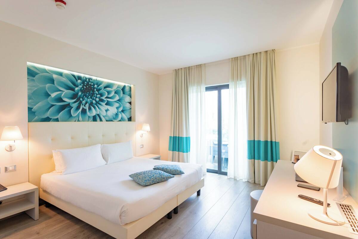 Une chambre d’hôtel avec un grand lit, des lampes de chevet, un bureau avec une lampe, une télévision et une grande œuvre d’art florale accrochée au mur.