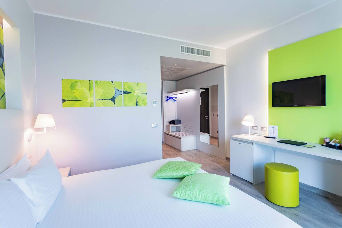Une chambre d’hôtel moderne avec un lit, des oreillers verts, un bureau avec une télévision et des œuvres d’art accrochées aux murs.