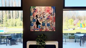 Una habitación con un cuadro abstracto grande en la pared, una planta en maceta y vistas a un espacio exterior con mesas y sillas.