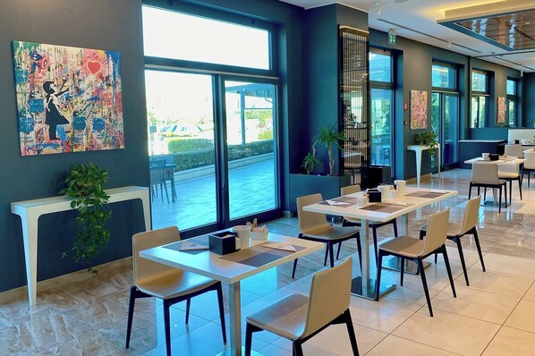 Un espace de restauration moderne, doté de grandes fenêtres, offrant une vue sur l’extérieur et agrémenté d’un tableau abstrait aux couleurs vives sur le mur.